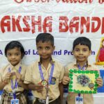 Three young students in beige uniforms pose with medals in front of a raksha bandhan banner at a school event